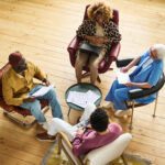 High angle of four intercultural economists having discussion of data at working meeting while sitting in armchairs in office with wooden floor
